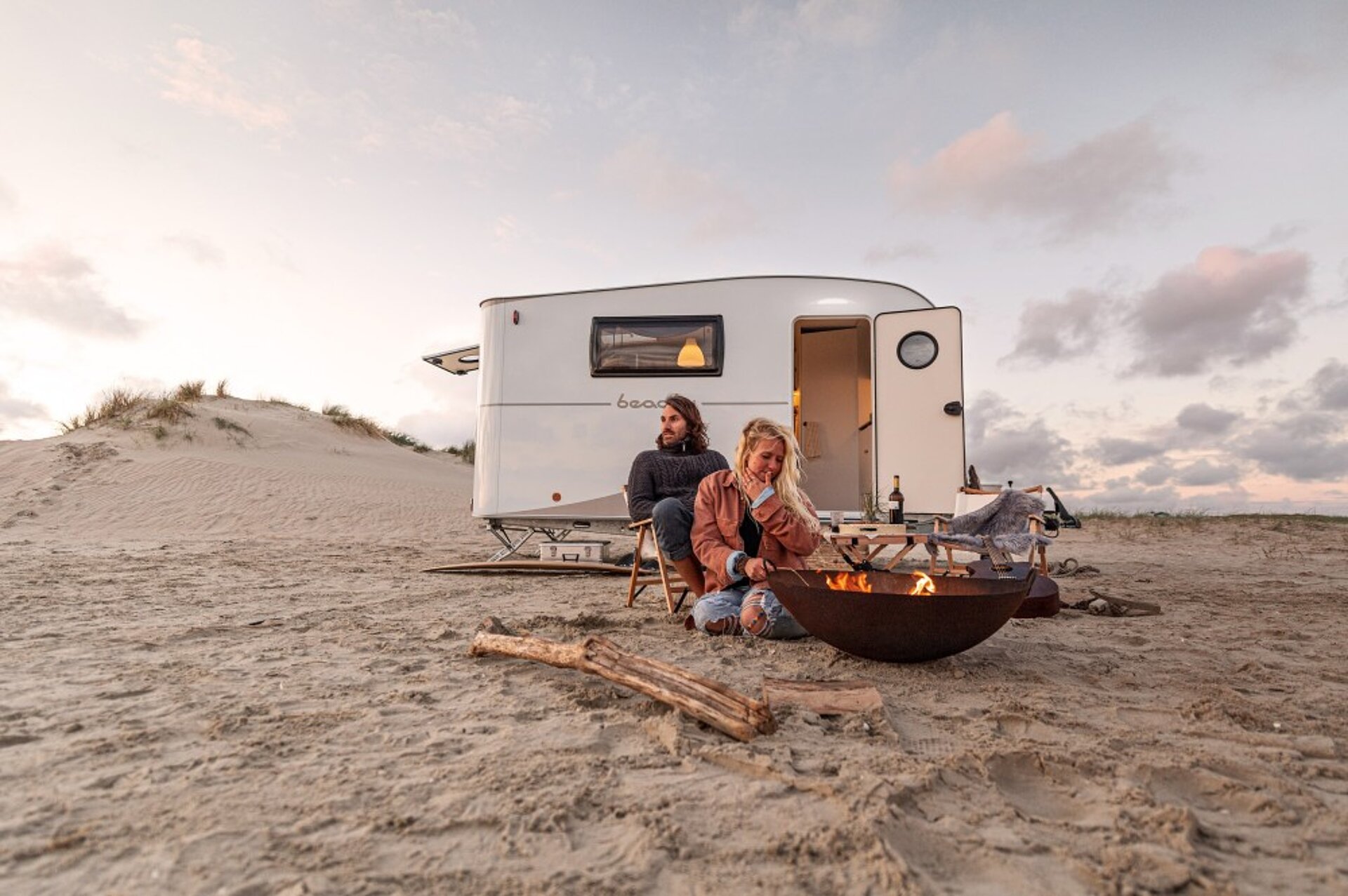 Ein Paar sitzt vor einem BEACHY Wohnwagen an einer Feuerschale am Strand.