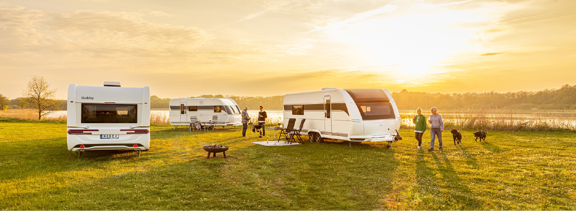 Entdecken Sie Ihr Freizeitfahrzeug bei Caravan Brandl! Mehrere Wohnwagen stehen auf einer Wiese, Menschen grillen davor.