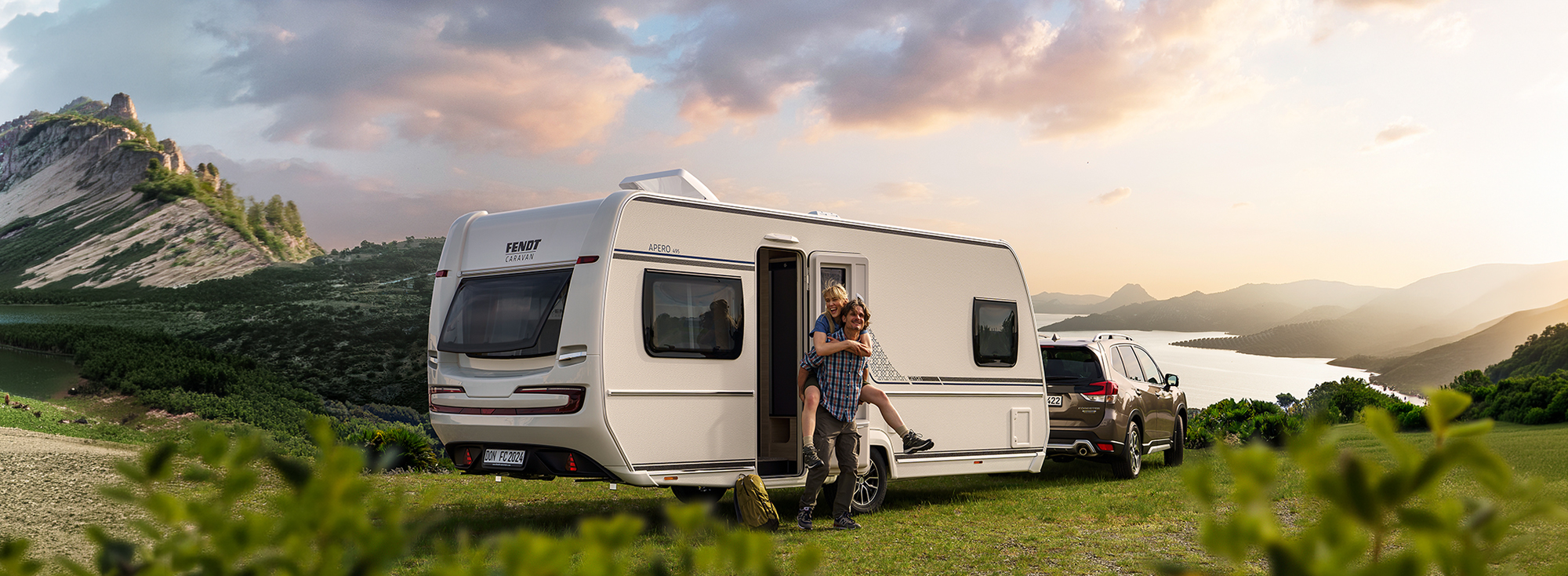 Ein Mann trägt seine Frau Huckepack vor einem geparkten Wohnwagen von Fendt-Caravan.